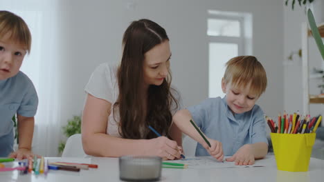 A-young-mother-with-two-children-sitting-at-a-white-table-draws-colored-pencils-on-paper-in-slow-motion